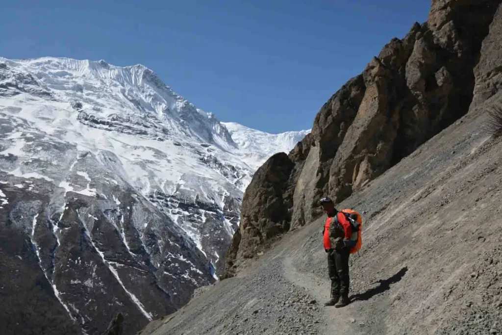 Trekking guide with an orange backpack cover standing on a mountain trail with snow-covered Himalayan peaks behind
